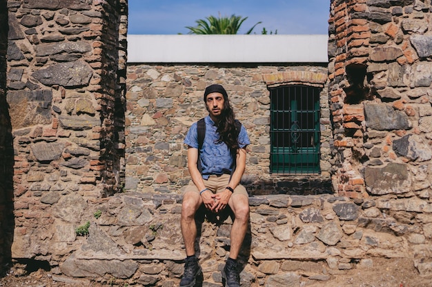 Giovane uomo latino con capelli lunghi che guarda l'obbiettivo seduto su un muro di ciottoli