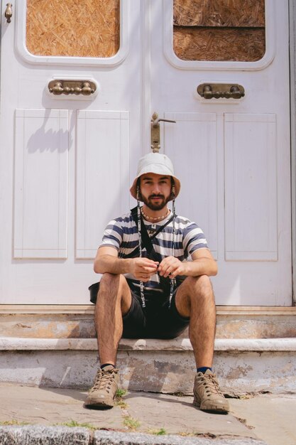 Giovane uomo latino con un cappello a secchiello seduto all'ingresso di una casa coloniale guardando la telecamera Foto verticale