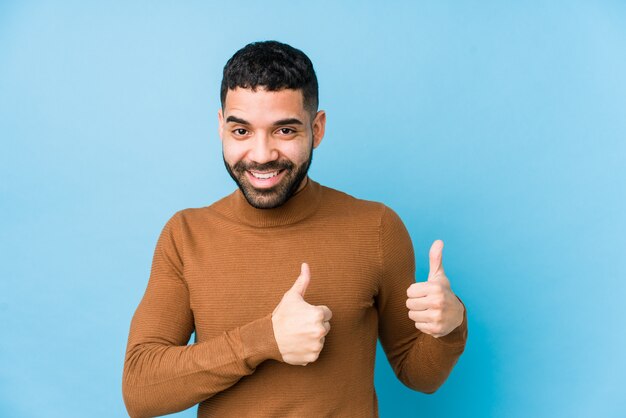 Giovane uomo latino contro una parete blu alzando entrambi i pollici, sorridendo e fiducioso.