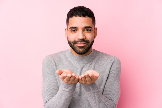 Giovane uomo latino contro una rosa isolato tenendo qualcosa con le palme, offrendo alla telecamera.