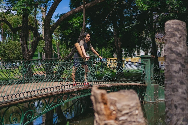 Giovane uomo latino hippie con i capelli lunghi appoggiato alla ringhiera di un ponte attraverso un lago che guarda verso l'orizzonte