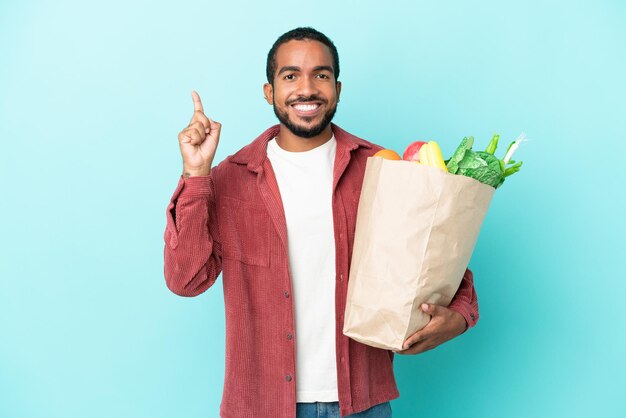 Giovane uomo latino in possesso di un sacchetto della spesa isolato su sfondo blu che indica una grande idea