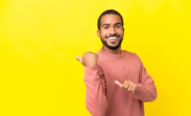 Giovane uomo latino isolato su sfondo giallo che indica il lato per presentare un prodotto