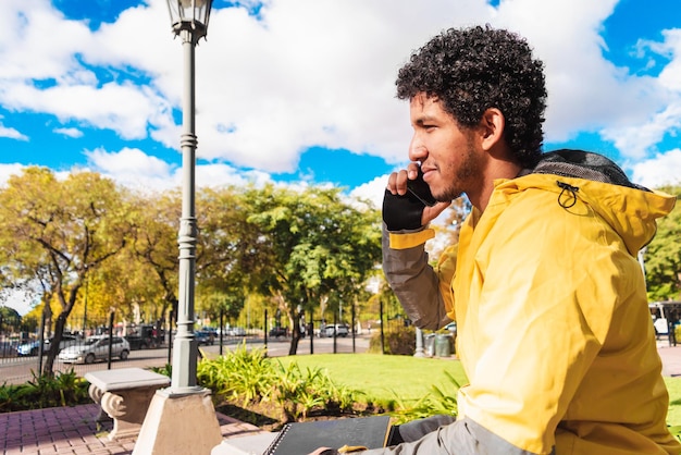Giovane uomo latino nero vestito di giallo seduto in una piazza urbana che parla al telefono