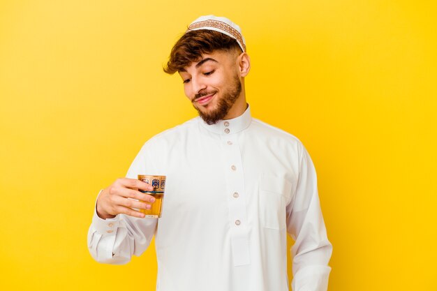 Giovane uomo marocchino che indossa il tipico costume arabo bere il tè isolato sulla parete gialla