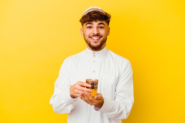 Giovane uomo marocchino che indossa il tipico costume arabo bere il tè isolato sulla parete gialla