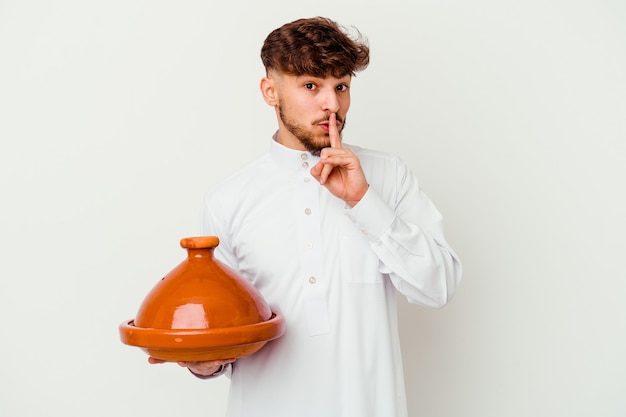 Giovane uomo marocchino che indossa il tipico costume arabo che tiene un tajine isolato su bianco mantenendo un segreto o chiedendo silenzio.