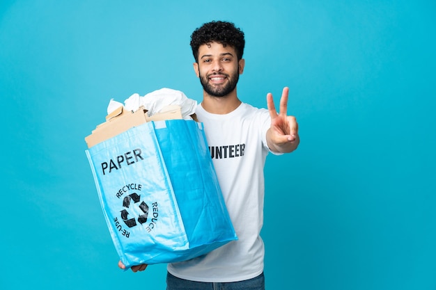 Giovane uomo marocchino che tiene un sacchetto di riciclaggio pieno di carta da riciclare sopra la parete isolata che sorride e che mostra il segno di vittoria
