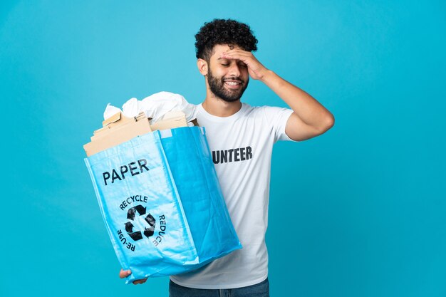 Giovane uomo marocchino che tiene un sacchetto di riciclaggio pieno di carta da riciclare sopra la parete isolata che sorride molto