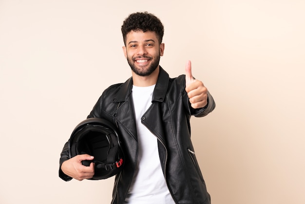 Giovane uomo marocchino con un casco da motociclista isolato sul muro beige con i pollici in su perché è successo qualcosa di buono