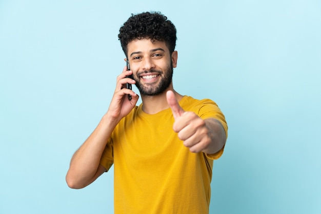 Giovane uomo marocchino isolato sulla parete blu mantenendo una conversazione con il cellulare mentre si fa i pollici in su