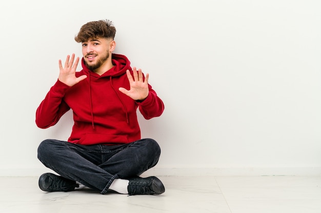 Giovane uomo marocchino seduto sul pavimento isolato su bianco rifiutando qualcuno che mostra un gesto di disgusto.