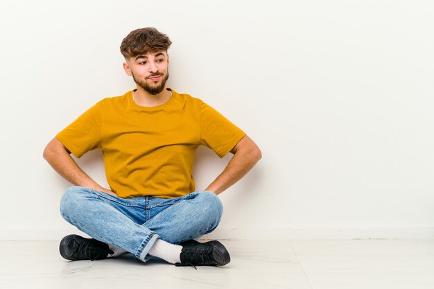 Giovane uomo marocchino seduto sul pavimento isolato su bianco sognando di raggiungere obiettivi e scopi