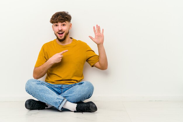 Giovane uomo marocchino seduto sul pavimento isolato su bianco sorridente allegro che mostra il numero cinque con le dita.