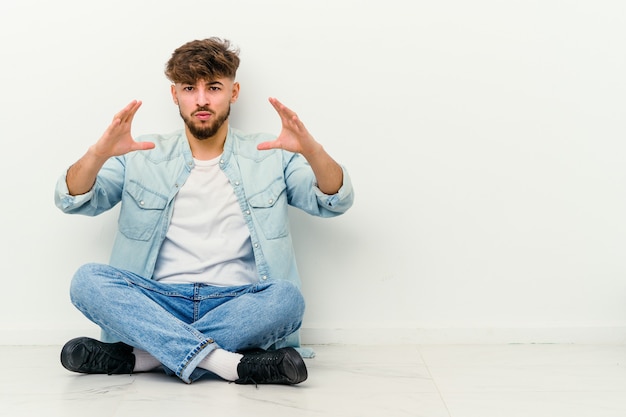 Giovane uomo marocchino seduto sul pavimento isolato su bianco tenendo qualcosa con le palme, offrendo alla telecamera.