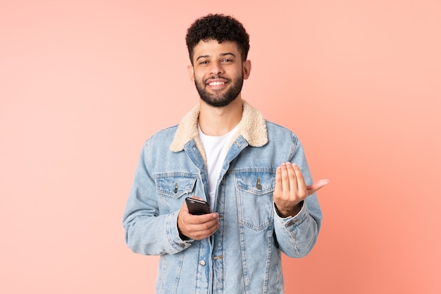 Giovane uomo marocchino utilizzando il telefono cellulare isolato sulla parete rosa che invita a venire con la mano. Felice che tu sia venuto