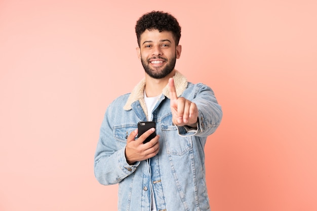 Giovane uomo marocchino utilizzando il telefono cellulare isolato sulla parete rosa che mostra e alzando un dito