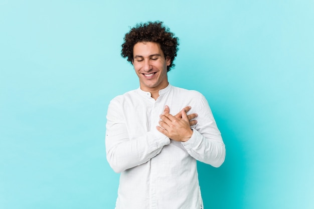 Giovane uomo maturo riccio che porta una camicia elegante che ride tenendo le mani sul cuore, concetto di felicità.