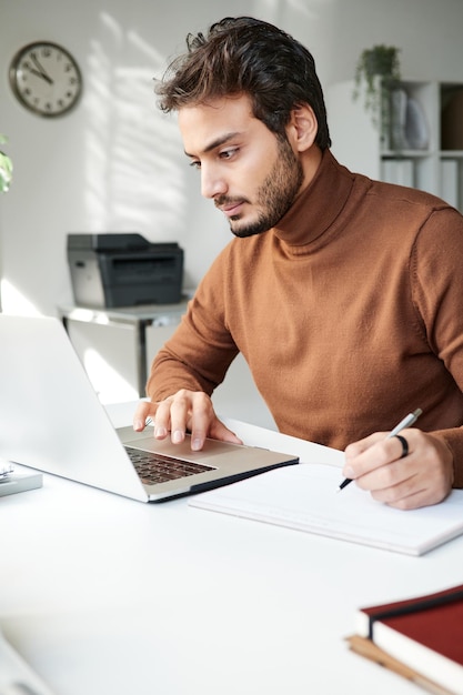 Giovane uomo mediorientale concentrato con stoppia seduto al tavolo e utilizzando il laptop mentre disegna uno schizzo in ufficio