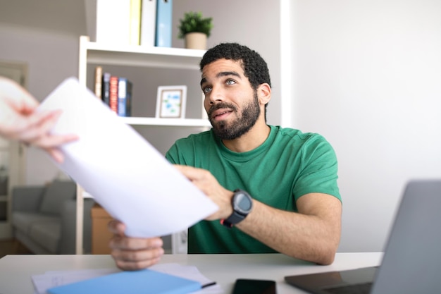 Giovane uomo mediorientale serio a tavola con il computer portatile che parla con il manager con i documenti