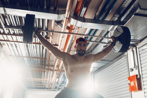 Giovane uomo muscoloso che fa esercizio di snatch o shoulder press con bilanciere su un duro allenamento presso la palestra del garage.