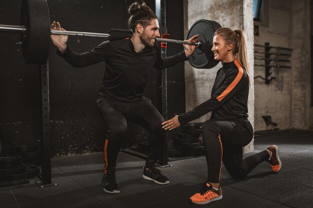 Giovane uomo muscoloso che fa esercizio tozzo con bilanciere in palestra con il supporto del suo personal trainer femminile.