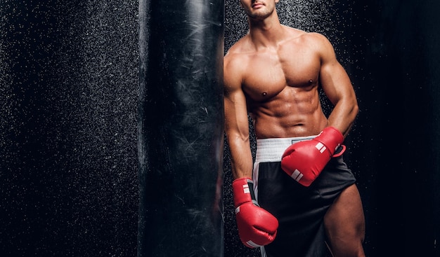 Giovane uomo muscoloso in guanti boxer rossi sta posando con un sacco da boxe in una stanza buia con spruzzi d'acqua sul muro.