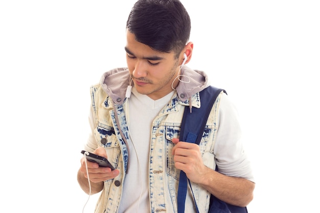 Giovane uomo ottimista in giacca di jeans con zaino blu utilizzando smartphone e cuffie in studio