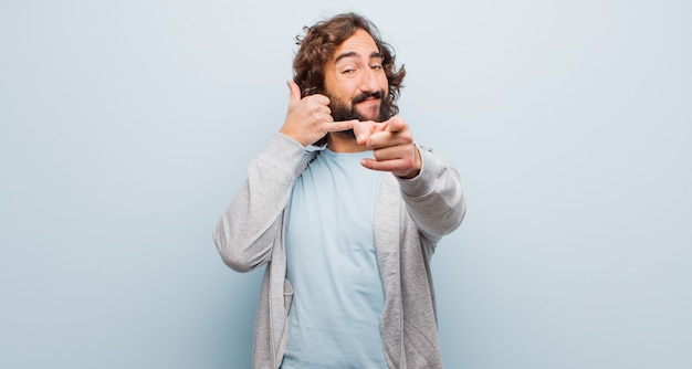 Giovane uomo pazzo barbuto che sorride allegramente e indicando mentre ti fa una chiamata in seguito gesto, parlando al telefono contro la parete di colore piatto