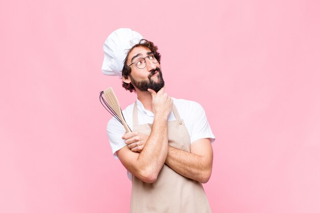Giovane uomo pazzo del panettiere con uno strumento cuoco contro il muro rosa