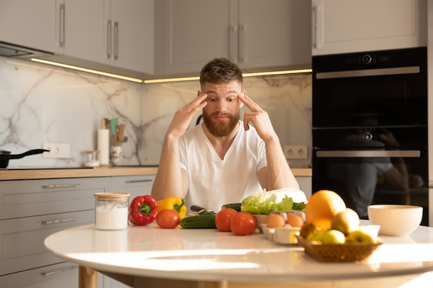 Giovane uomo preoccupato seduto a tavola con diversi ortaggi biologici e freschi, frutta, uova e sale. Ragazzo barbuto europeo. Interno della cucina in appartamento moderno. Concetto di alimentazione sana