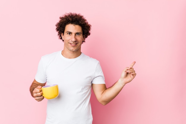 Giovane uomo riccio caucasico che tiene una tazza di tè che sorride allegramente indicando con l'indice di distanza.