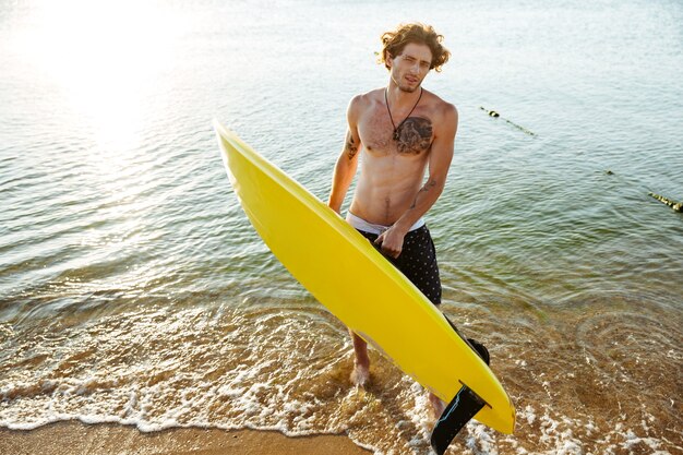 Giovane uomo riccio che tiene la sua tavola da surf mentre si trovava nell'oceano in spiaggia