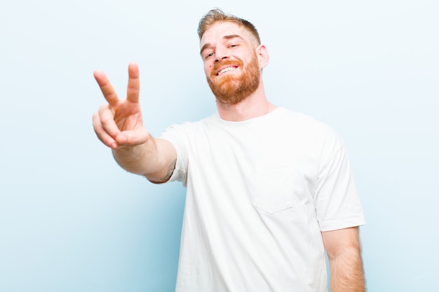 Giovane uomo rosso che sorride e che sembra vittoria felice, spensierata e positiva, gesturing o pace con una mano contro la parete blu molle