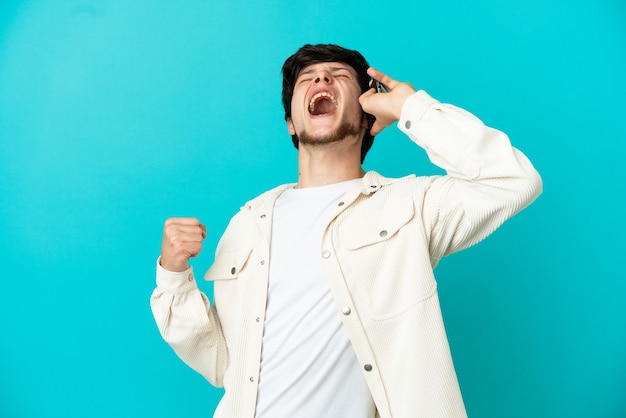 Giovane uomo russo che utilizza il telefono cellulare isolato su sfondo blu che celebra una vittoria