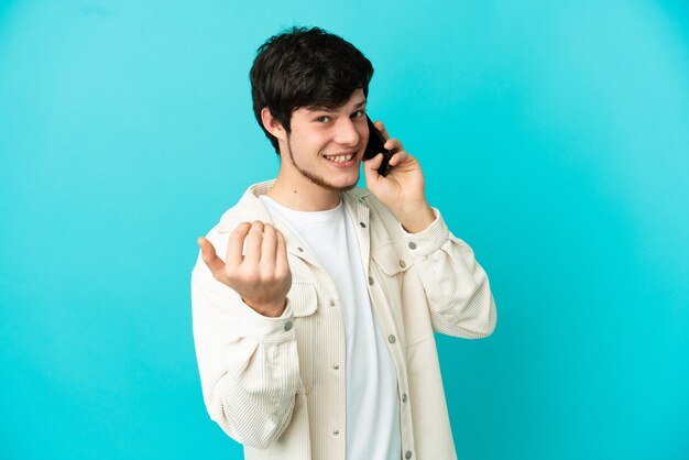 Giovane uomo russo che utilizza il telefono cellulare isolato su sfondo blu che invita a venire con la mano. Felice che tu sia venuto