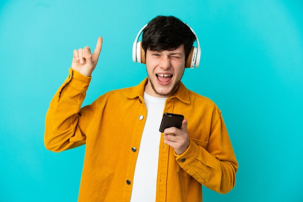 Giovane uomo russo isolato su sfondo blu ascoltando musica con un cellulare che fa gesto rock