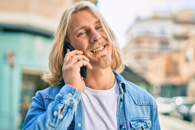 Giovane uomo scandinavo biondo che sorride felice parlando sullo smartphone in città