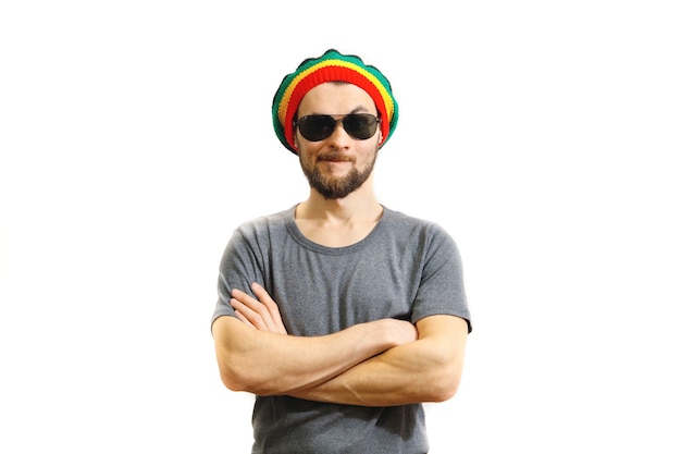 Giovane uomo scettico caucasico in cappello rasta, occhiali da sole e maglietta grigia su sfondo bianco.