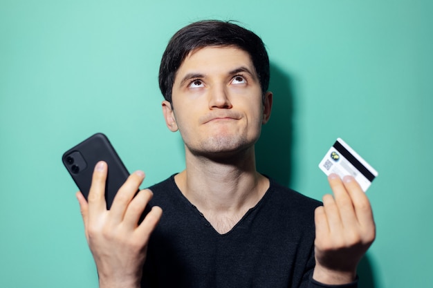giovane uomo sconvolto cercando tenendo in mano lo smartphone e la carta di credito sul muro di colore aqua menthe.