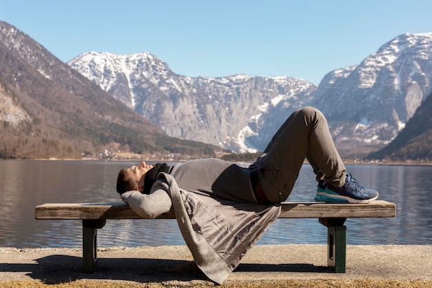 Giovane uomo sdraiato all'aperto su una panchina e godersi le montagne neve bel tempo cielo blu sole Bellissimo paesaggio Tempo con te stesso sognando relax salute mentale Turismo vacanze viaggi