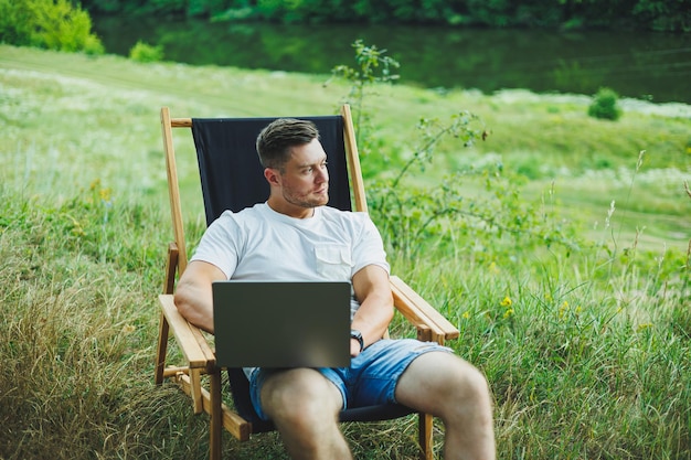 Giovane uomo sdraiato su una sedia nella natura e che lavora con un computer portatile riposando da solo guardando pensieroso Un uomo in viaggio in campagna il concetto di stile di vita delle persone