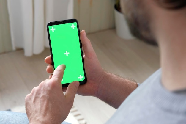 Giovane uomo seduto a casa tenendo in mano lo schermo mockup verde dello smartphone Persona di sesso maschile che utilizza il telefono cellulare chroma key Modalità verticale Toccando lo scorrimento del display toccando la navigazione su Internet social media