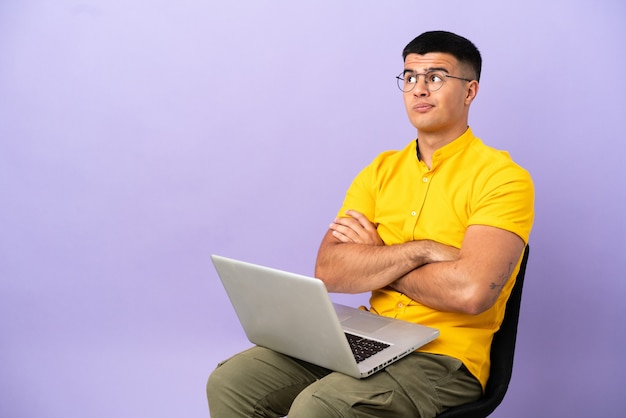 Giovane uomo seduto su una sedia con il computer portatile che fa gesti di dubbio mentre solleva le spalle