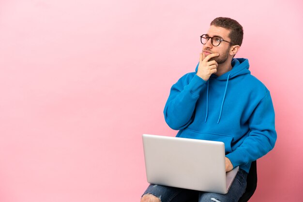 Giovane uomo seduto su una sedia con il computer portatile che ha dubbi e con un'espressione del viso confusa