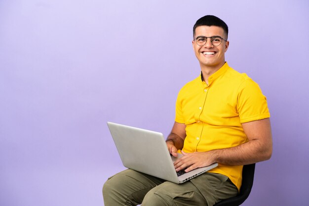 Giovane uomo seduto su una sedia con il computer portatile in posa con le braccia all'anca e sorridente