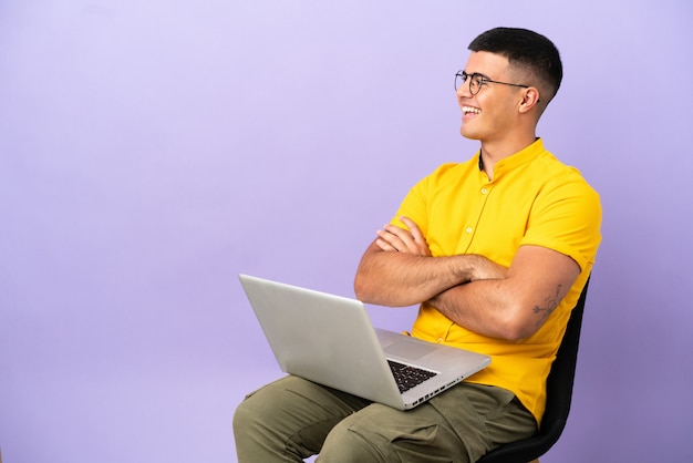 Giovane uomo seduto su una sedia con laptop felice e sorridente happy