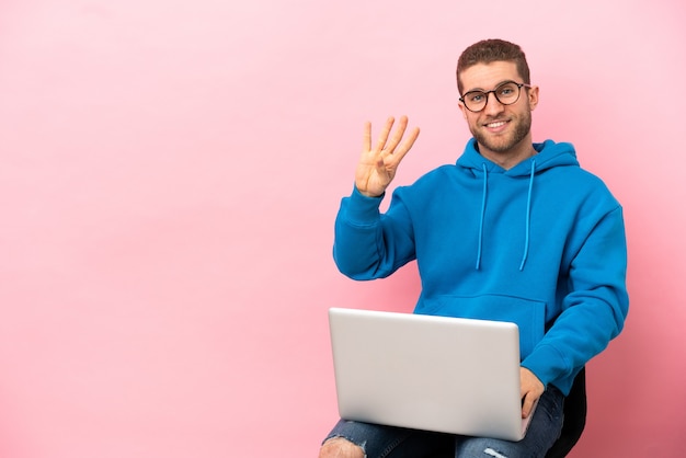 Giovane uomo seduto su una sedia con un laptop felice e che conta quattro con le dita