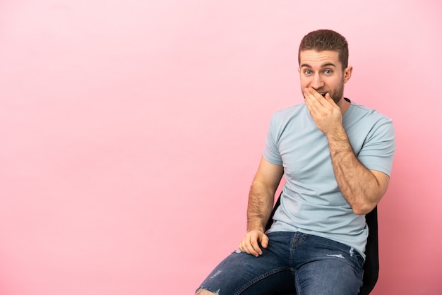 Giovane uomo seduto su una sedia sopra isolato felice e sorridente che copre la bocca con la mano