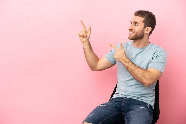 Giovane uomo seduto su una sedia su sfondo rosa isolato che indica con il dito indice una grande idea